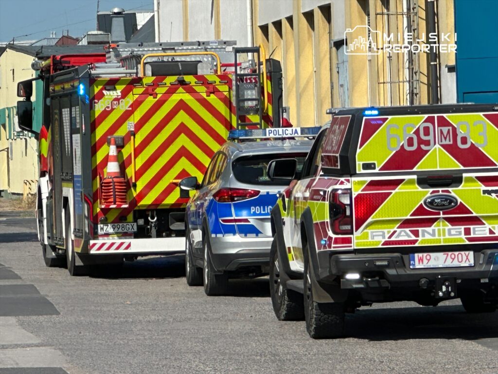 Wozu strażacki z migającymi światłami parkuje obok radiowozu policyjnego na wąskiej ulicy.