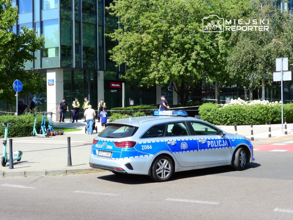 Policyjny radiowóz z niebieskimi lampami stoi na ulicy, a obok grupka funkcjonariuszy rozmawia z przechodniami.