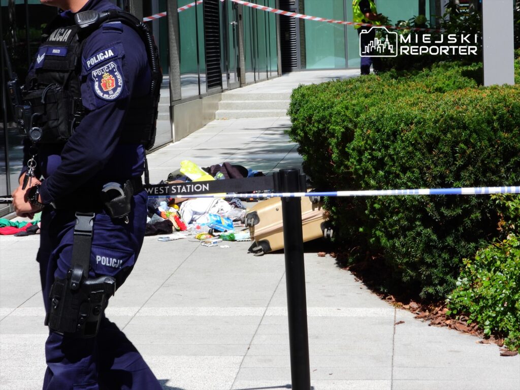 Policjant w mundurze stoi przy taśmie zabezpieczającej, obok leżą różne przedmioty i walizka.