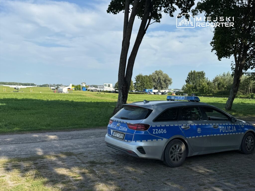 Policyjny samochód zaparkowany na drodze w pobliżu lotniska, w tle widoczne samoloty i zielona trawa.