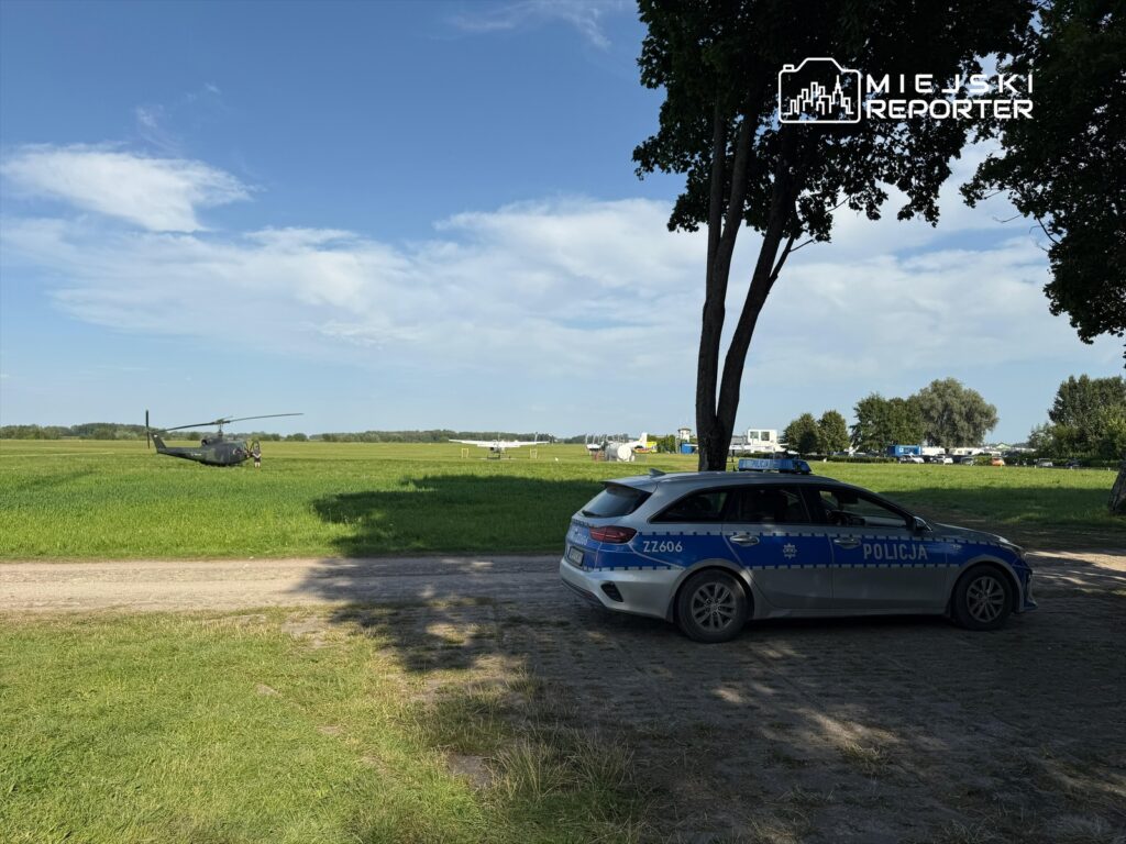 Helikopter wojskowy ląduje na zielonym polu, obok stoi radiowóz policji. W tle widoczne inne pojazdy.