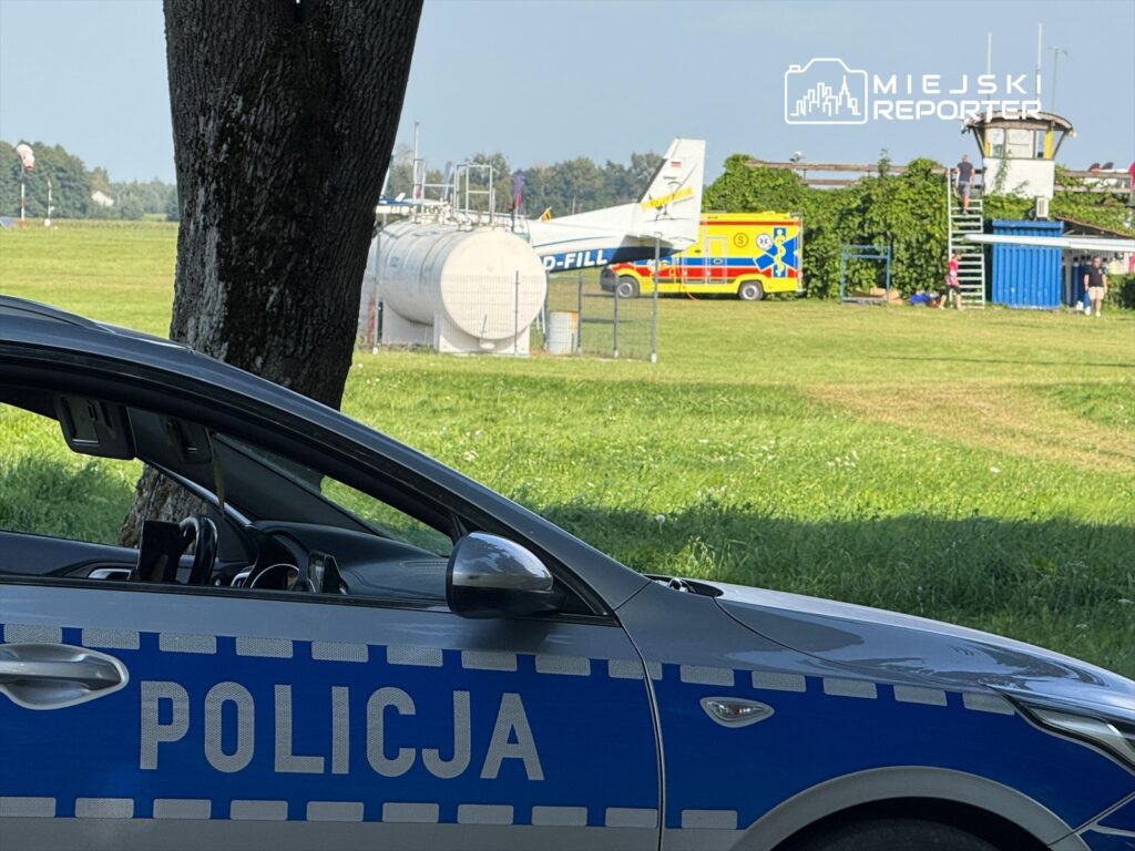 Policyjny samochód zaparkowany w pobliżu lądowiska, w tle karetka pogotowia i hangar lotniczy.