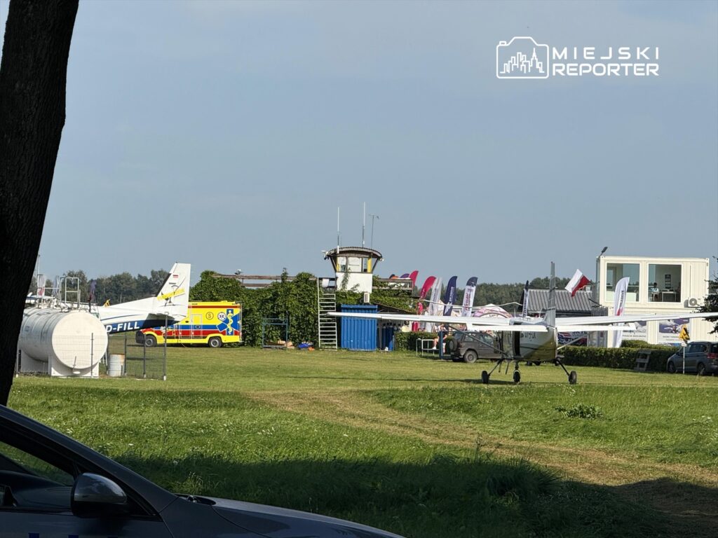 Mały samolot na płycie lotniska, obok karetka i wieża kontrolna, w tle budynki z flagami.