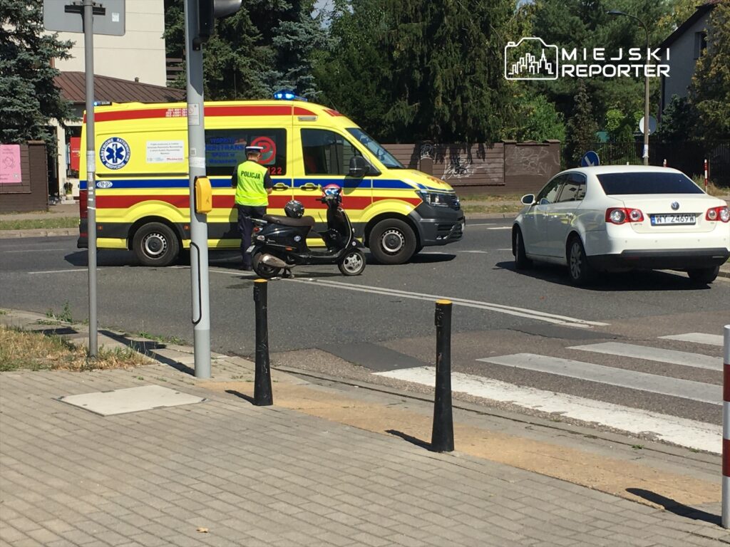 Karetka pogotowia z włączonymi sygnałami świetlnymi zatrzymana na skrzyżowaniu, obok policjant na skuterze.