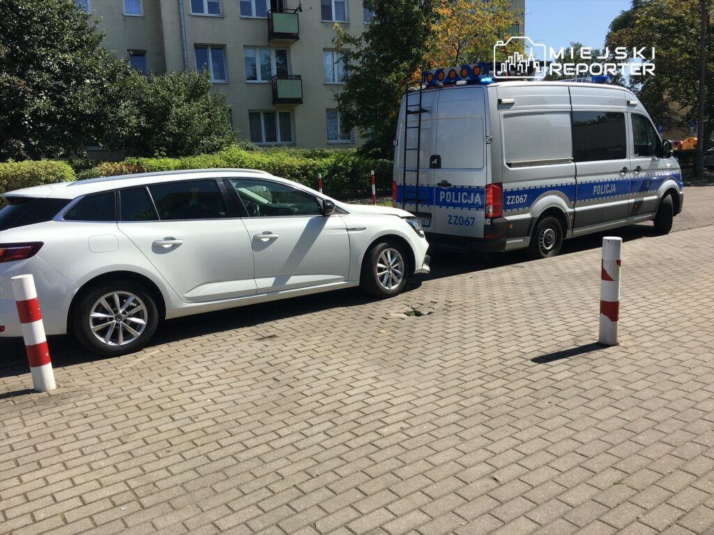 Biały samochód osobowy zaparkowany obok policyjnego vana na parkingu, w tle budynki mieszkalne.