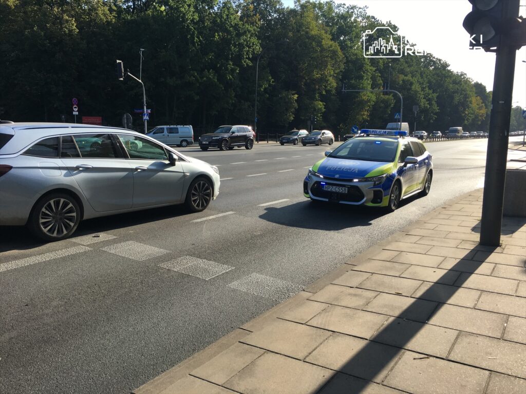 Policyjny radiowóz z włączonymi sygnałami świetlnymi zatrzymuje się na skrzyżowaniu, obok innych samochodów.