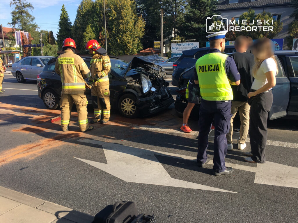 Strażacy w mundurach, zjeżdżający do wypadku samochodowego, rozmawiają z policjantem i osobami poszkodowanymi na ruchliwej ulicy.