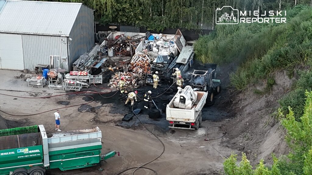 Grupa strażaków gasi pożar w pobliżu składowiska odpadów, obok uszkodzonego pojazdu ciężarowego.
