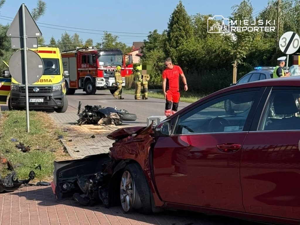Czerwony samochód osobowy uszkodzony po wypadku, w tle strażacy i karetka na miejscu zdarzenia.