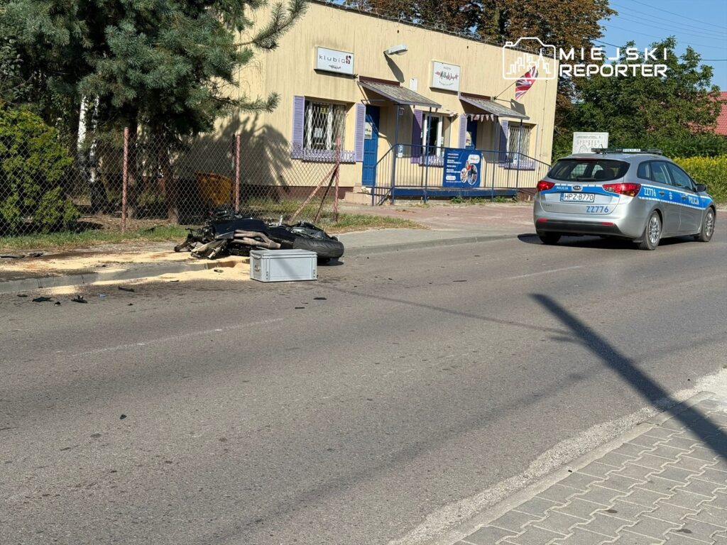 Na jezdni leży uszkodzony motocykl oraz rozrzucone fragmenty, obok przejeżdża radiowóz policyjny.