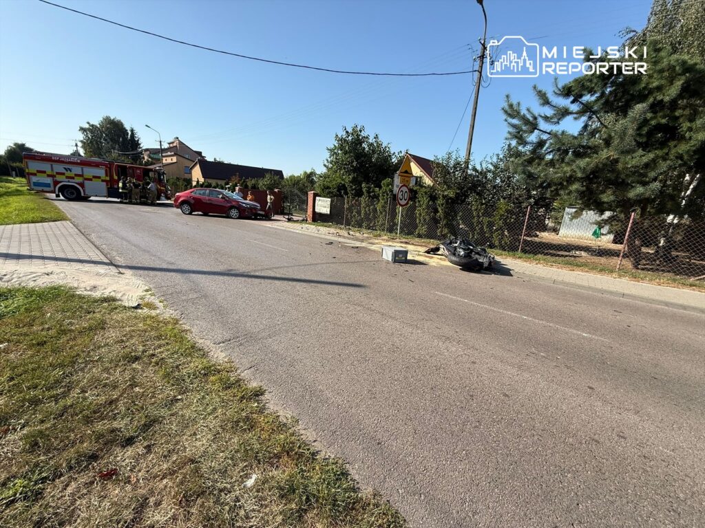 Na drodze stoi czerwona osobówka, a obok niej zespół strażaków przy wozie strażackim. W tle widoczny zniszczony motocykl.