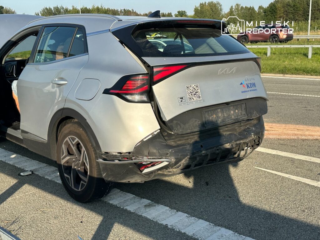 Uszkodzony srebrny samochód SUV stojący na poboczu drogi, z otwartymi drzwiami i widocznymi uszkodzeniami tylnej części.