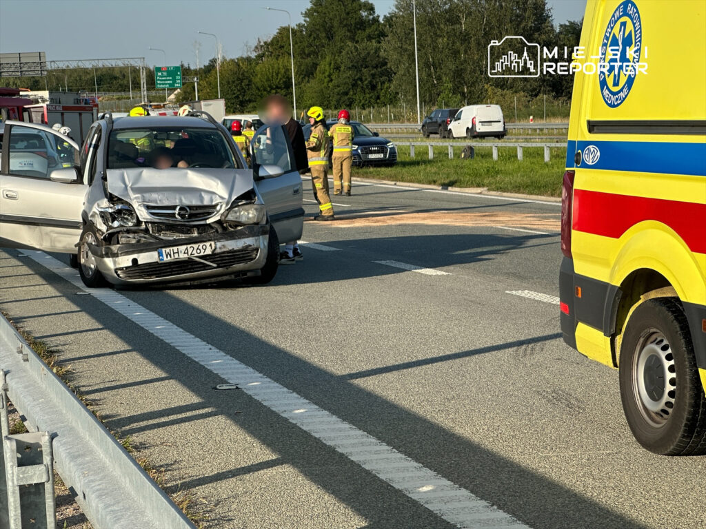 Uszkodzony samochód osobowy z wgniecioną maską stoi na pasie ruchu, obok grupy ratowników medycznych oraz strażaków.