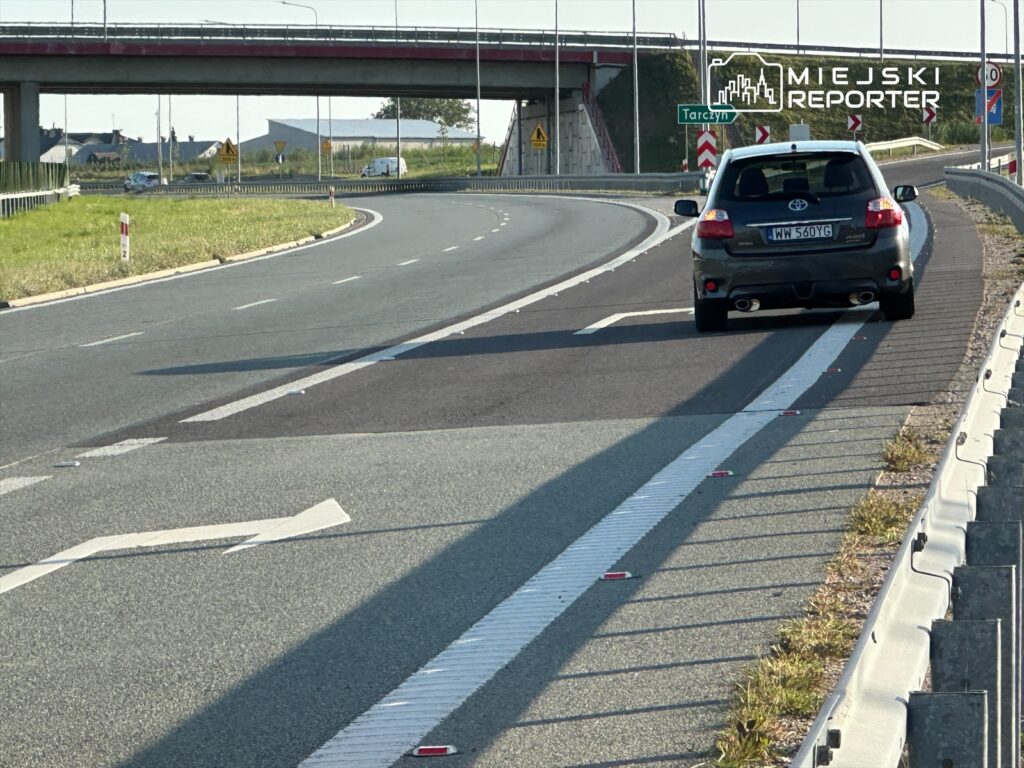 Samochód osobowy zjeżdża z drogi na łuku, obok znaku kierunkowym wskazującym na miejscowość Tarczyn.