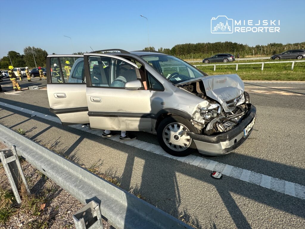 Uszkodzony samochód osobowy z wgniecionym przodem stoi na jezdni, obok pracują strażacy oraz służby ratunkowe.