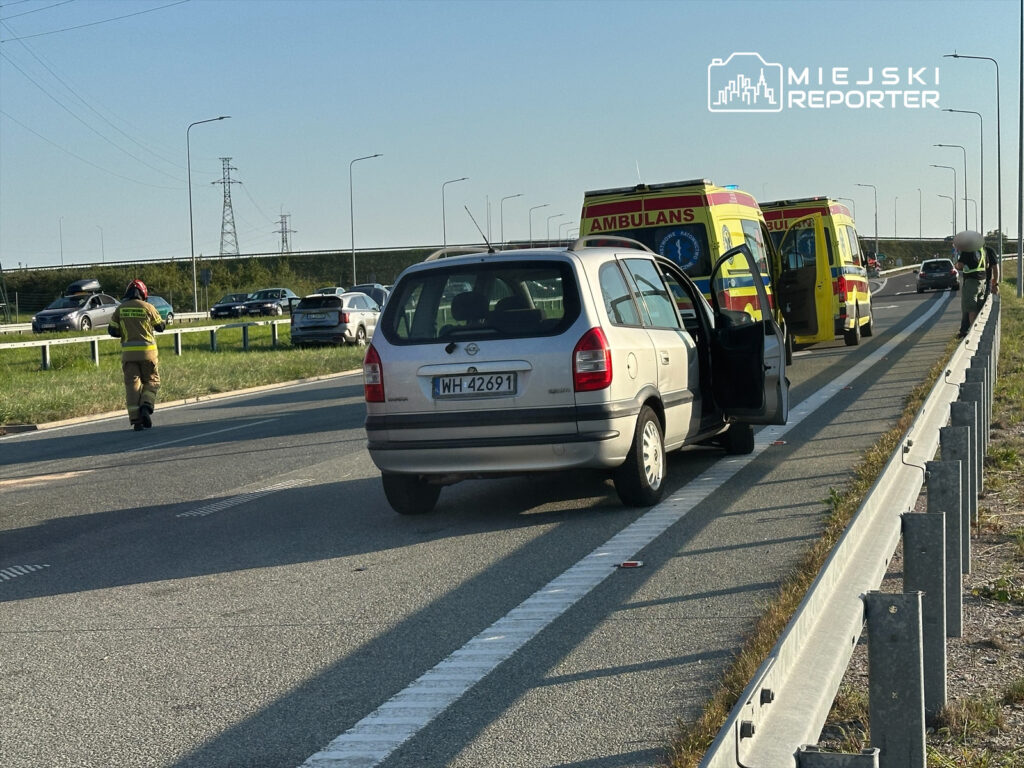 Kierowca w mundurze ratunkowym idzie w stronę stojącego samochodu osobowego przy drodze, obok parkujących ambulansów.