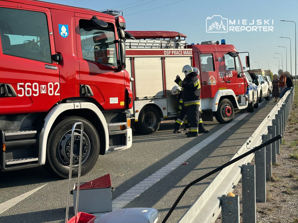 Strażacy w mundurach rozmawiają przy dwóch wozach strażackich na poboczu drogi, obok zatrzymanych pojazdów.