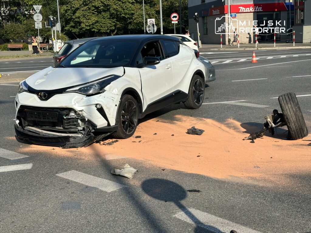 Uszkodzony biały samochód osobowy stoi na skrzyżowaniu, obok leżący oderwany tylni koło na drodze.