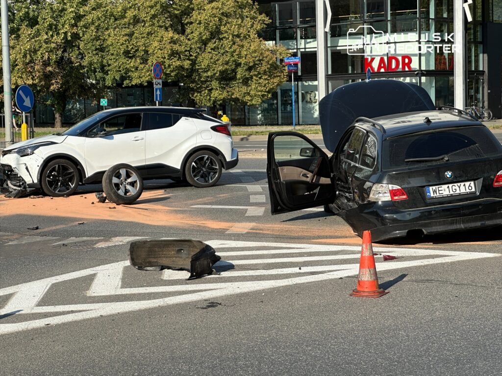 Dwa uszkodzone samochody stoją na rondzie, jeden z otwartymi drzwiami, wokół leżą fragmenty pojazdów i pachołek ostrzegawczy.