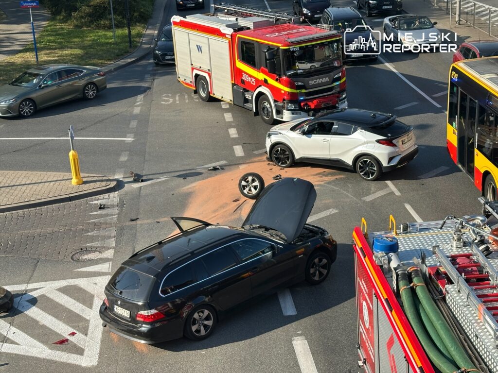 Czarny samochód osobowy z otwartą maską stoi na skrzyżowaniu obok wozu strażackiego, a w tle widoczne inne pojazdy.