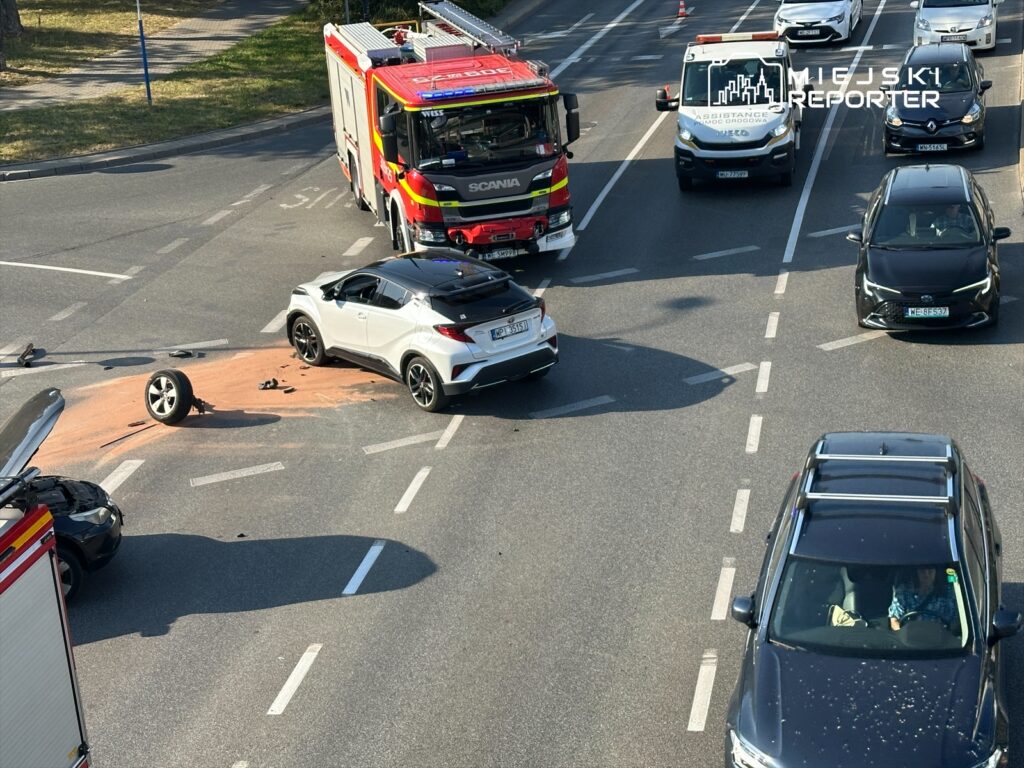 Zderzenie dwóch samochodów na skrzyżowaniu, z białym pojazdem uszkodzonym i leżącym kołem na jezdni. W tle widać wóz strażacki i ambulans.