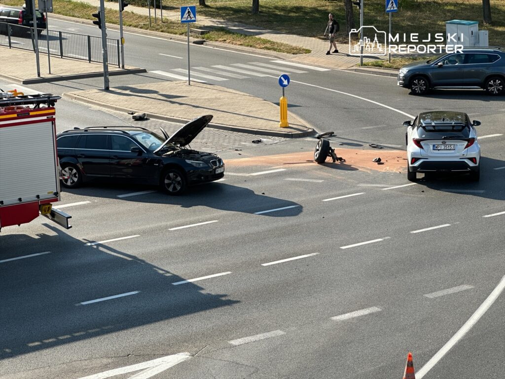 Dwa uszkodzone pojazdy na skrzyżowaniu, jeden z otwartą maską, a obok leżący fragment motocykla.