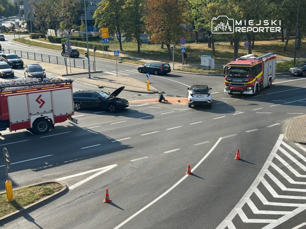 Samochód osobowy z otwartą maską stoi na skrzyżowaniu obok wozu strażackiego, na drodze stoją pachołki.