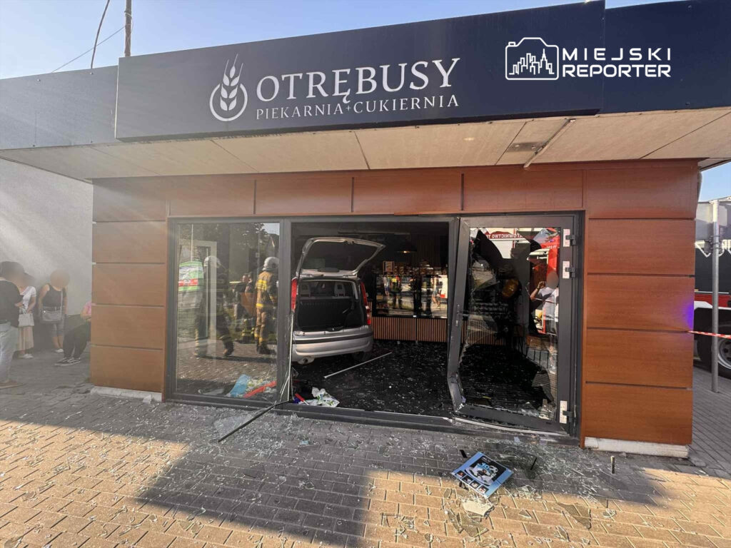 Samochód wjechał w witrynę piekarni, a wokół leżą odłamki szkła i porozrzucane przedmioty. W tle widać osoby obserwujące zdarzenie.