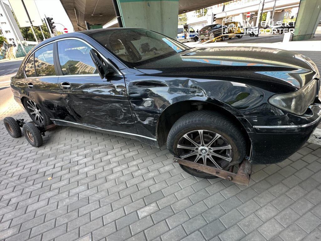 Czarny samochód osobowy z uszkodzoną karoserią stoi na chodniku, na dwóch oponach, z przymocowanymi wózkami.
