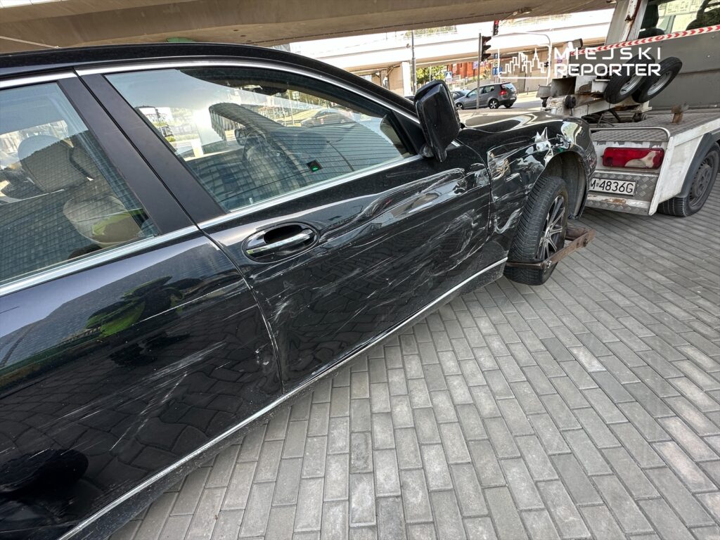 Czarny samochód osobowy z uszkodzeniami na prawym boku stoi na płycie chodnikowej obok lawety.