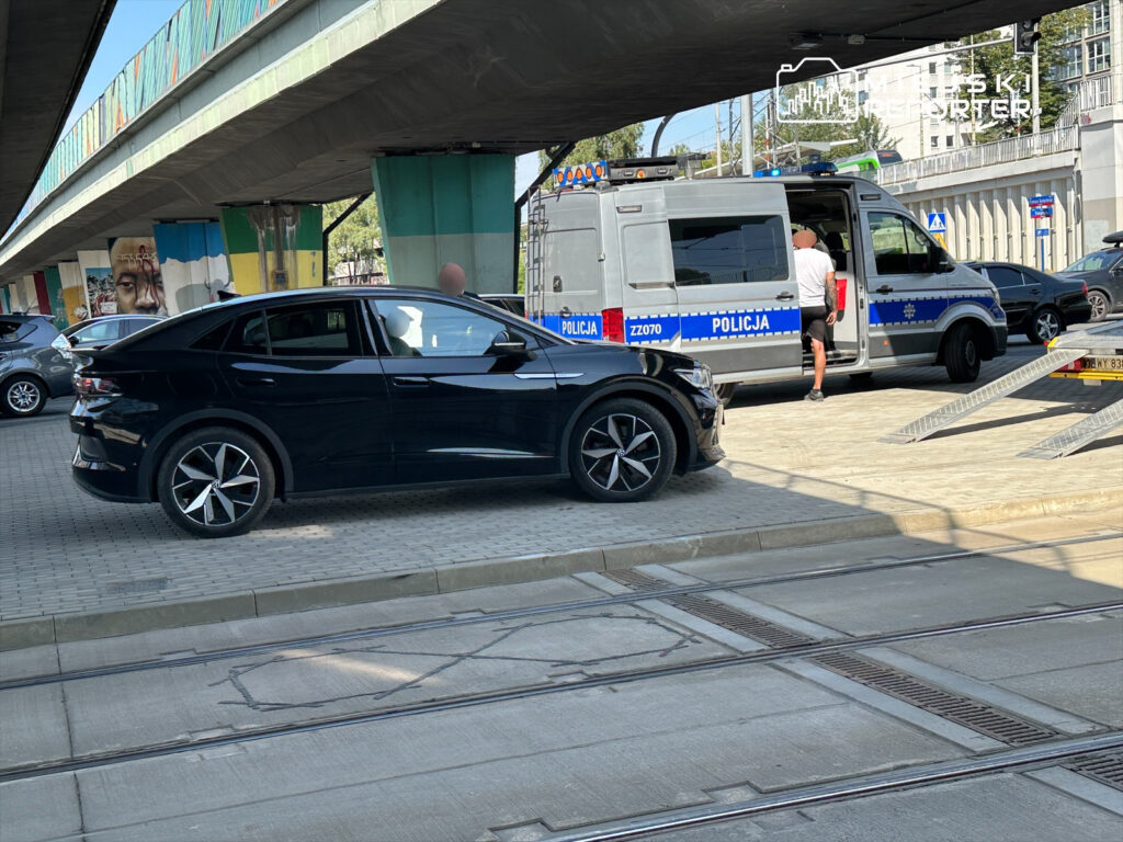 Czarny samochód osobowy zaparkowany obok radiowozu policyjnego, w tle widoczne graffiti na murze.