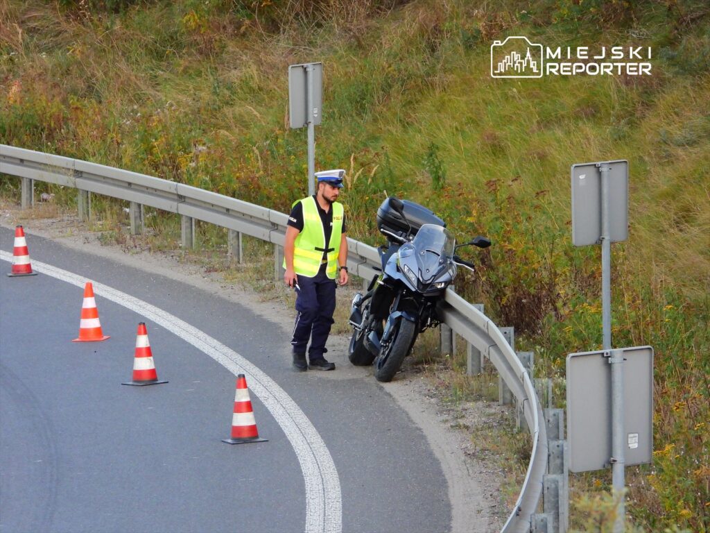 Policjant w odblaskowej kamizelce kontroluje motocykl zaparkowany przy drodze, otoczony pachołkami.