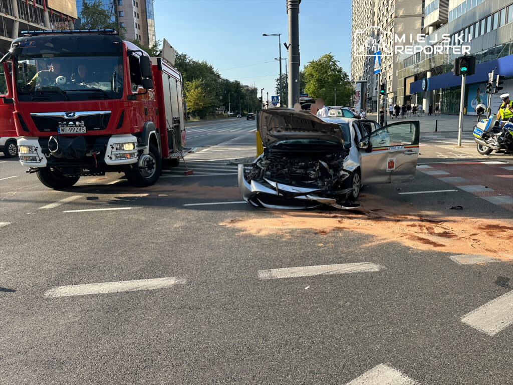 Czerwony wóz strażacki zatrzymany obok uszkodzonego samochodu osobowego na ruchliwej ulicy, wokół widoczni policjanci.