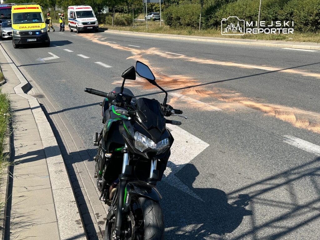 Motocykl stoi na poboczu drogi, obok widać karetki i służby ratunkowe, a na jezdni widoczne są plamy.