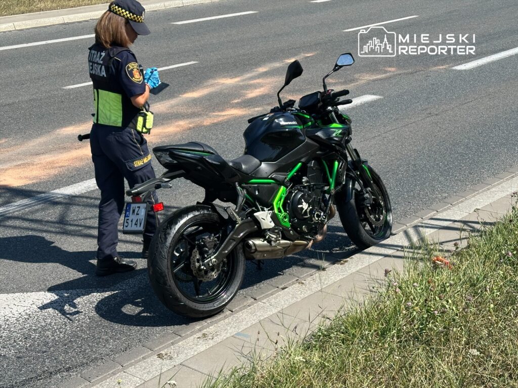 Kobieta w mundurze straży miejskiej notuje informacje obok motocykla na poboczu drogi.