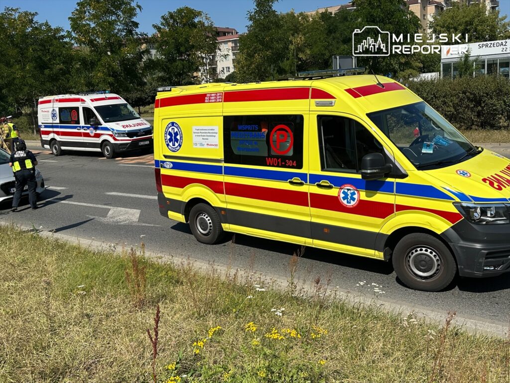 Karetka pogotowia ratunkowego zatrzymana na drodze, obok policyjny radiowóz i druga karetka w tle.
