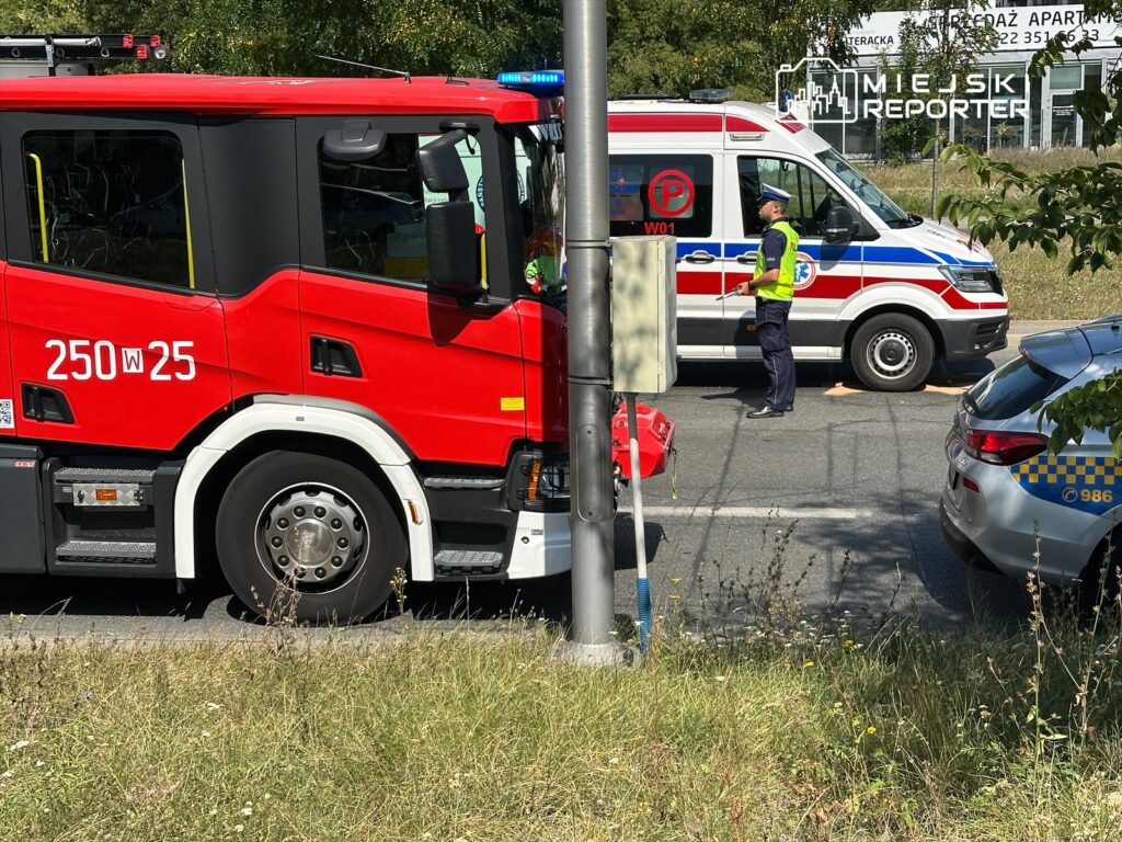 Czerwony wóz strażacki stoi obok karetki pogotowia, a policjant kieruje ruchem na drodze.