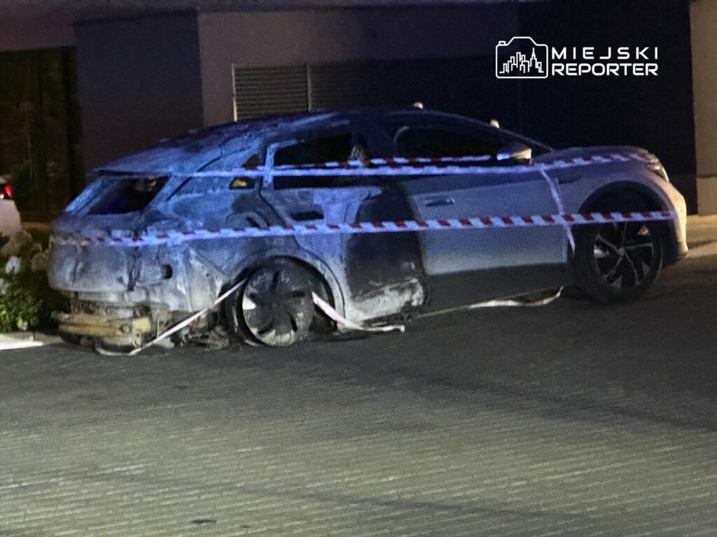 Spalony samochód osobowy zabezpieczony taśmą policyjną na parkingu przy nowoczesnym budynku.