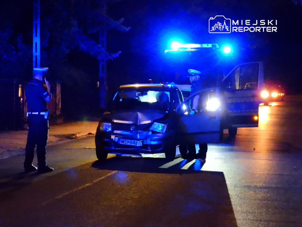Policjant rozmawia przez telefon obok uszkodzonego samochodu na oświetlonej ulicy nocą. W tle widoczny radiowóz.