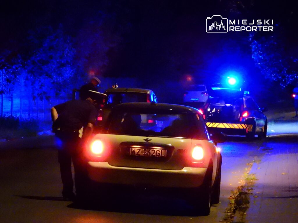 Policjant kontrolujący kierowcę w małym samochodzie na ulicy oświetlonej niebieskimi światłami radiowozu.