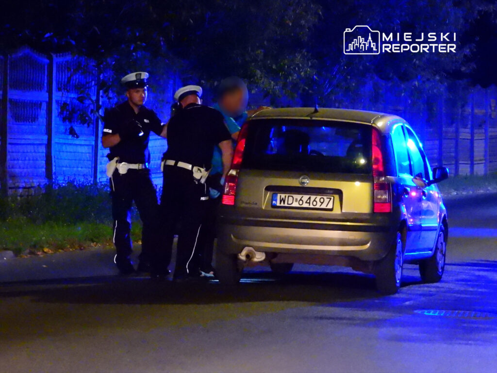 Dwaj policjanci kontrolują mężczyznę przy zaparkowanym samochodzie w nocy, w tle niebieskie światła.