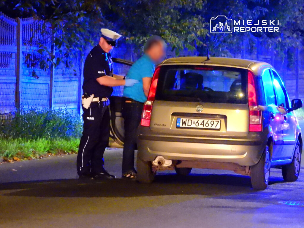 Policjant w mundurze kontroluje mężczyznę przy otwartym bagażniku samochodu osobowego w nocy.