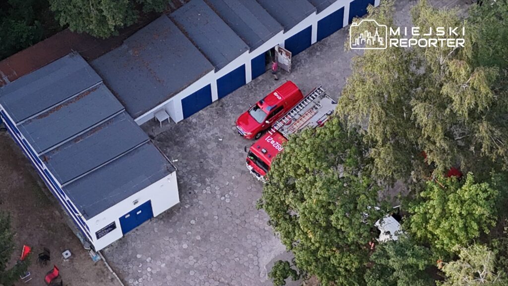 Czerwony wóz strażacki zaparkowany obok garaży, obok drabina strażacka, w otoczeniu drzew.