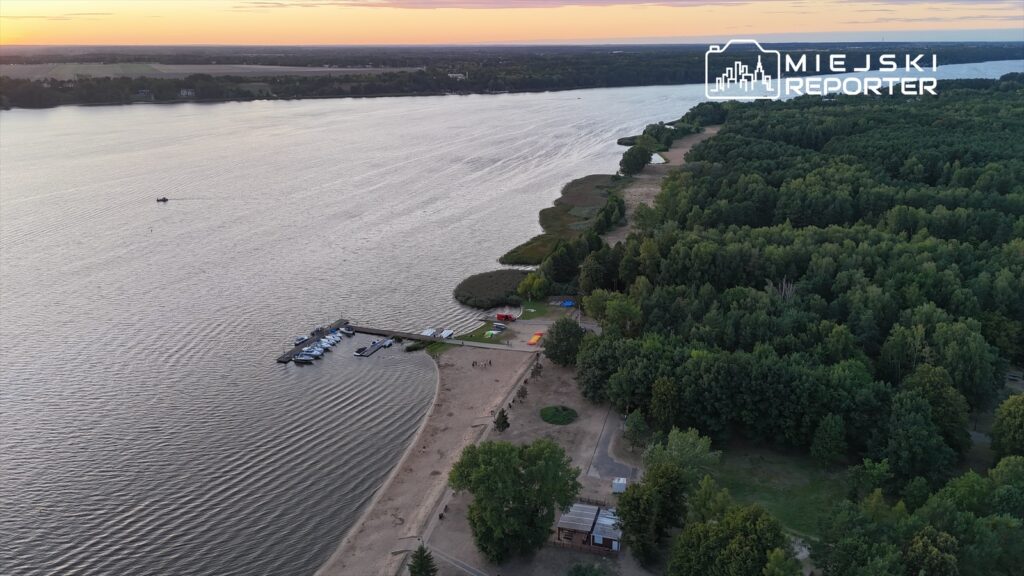 Widok na rzekę z przystanią, gdzie cumują łodzie, w otoczeniu zielonych drzew i piaszczystej plaży.