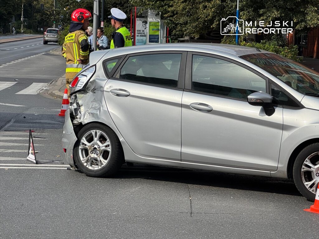 Kierowca w mundurze rozmawia z ratownikiem w kasku obok uszkodzonego samochodu na drodze.