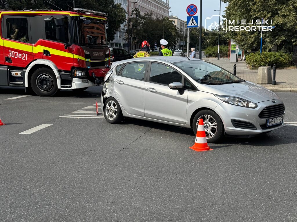 Wypadek drogowy z udziałem srebrnego samochodu osobowego oraz wozu strażackiego na skrzyżowaniu, otoczenie miejskie.