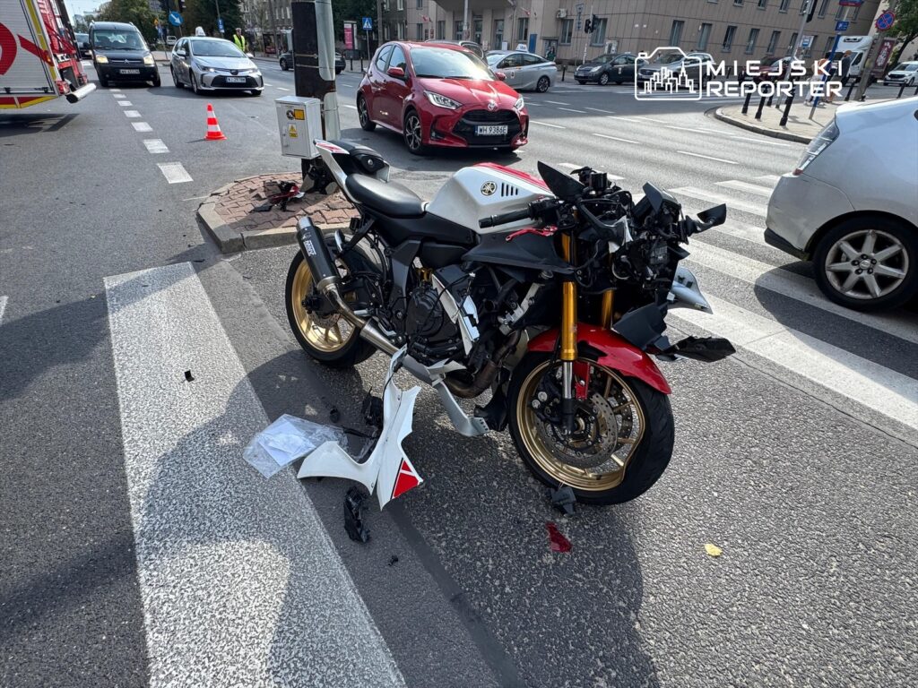 Motocykl z uszkodzoną przednią częścią stoi na jezdni, obok ruchliwego skrzyżowania z samochodami w tle.