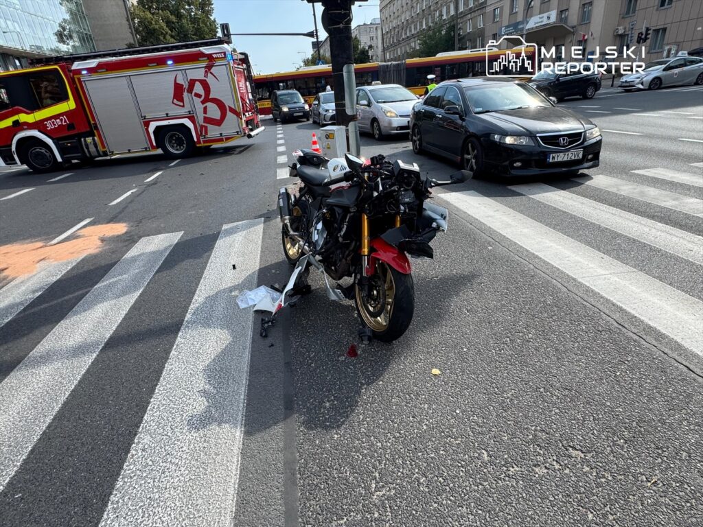 Motocykl z uszkodzeniami leży na przejściu dla pieszych, obok widać wóz strażacki i ruch uliczny.