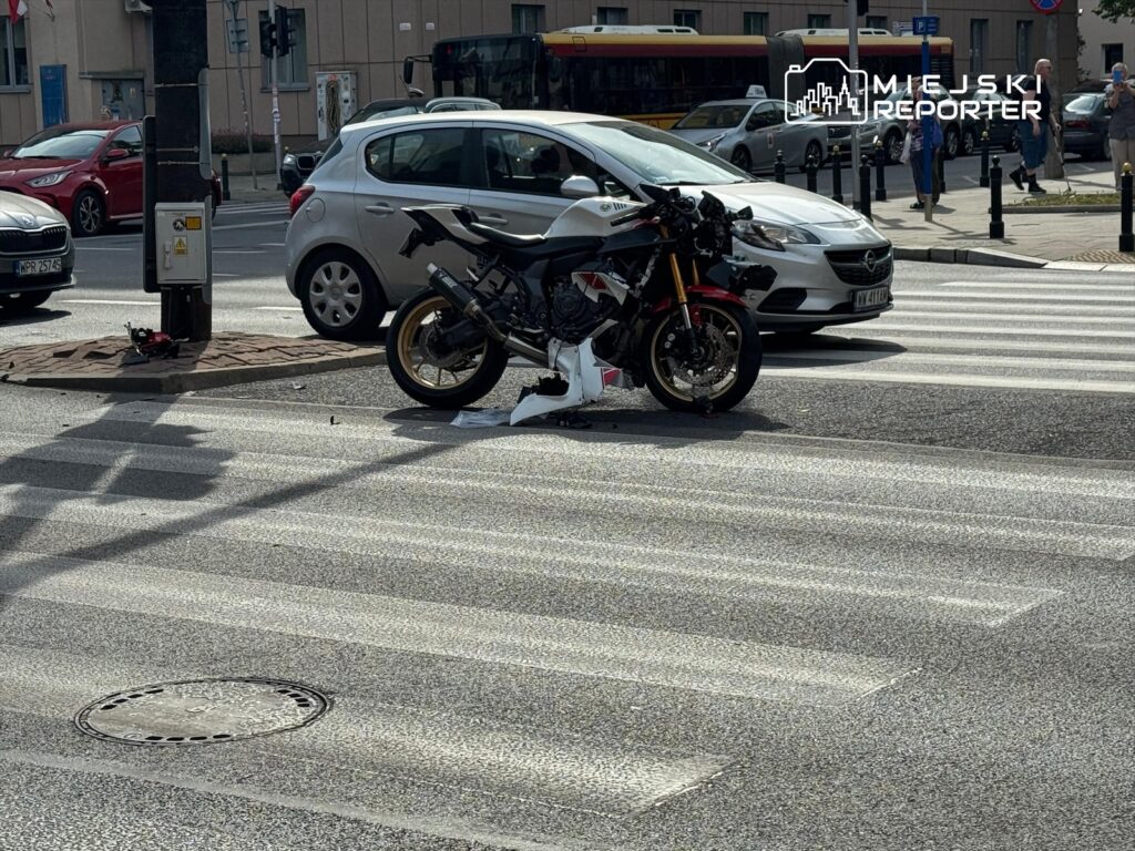 Motocykl leży na pasach, obok uszkodzonego samochodu osobowego, w miejskim otoczeniu.