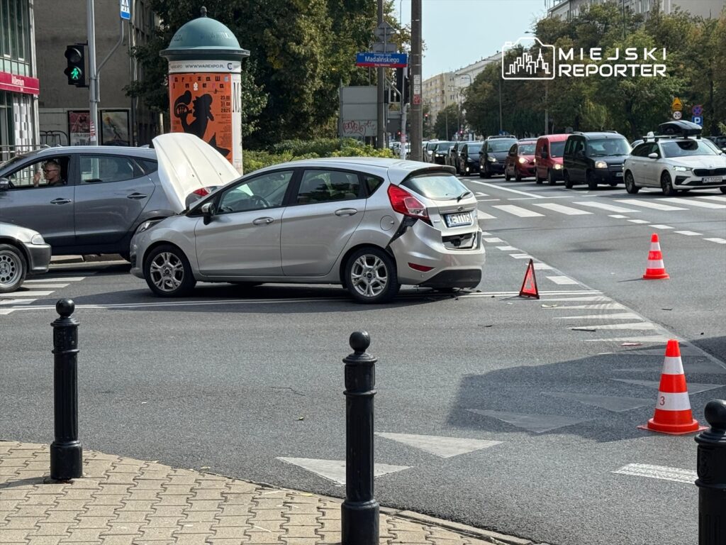 Dwa samochody zderzone na skrzyżowaniu, jeden z otwartą maską, wokół trójkąty ostrzegawcze. Ruch uliczny spowolniony.
